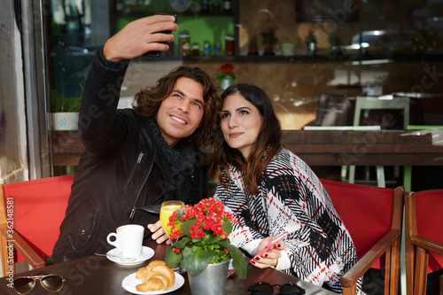 Couple in love breakfast with warm coffee and delicious dessert in sidewalk cafe  attractive man and woman taking picture of themselves with mobile phone camera during vacation holidays in autumn