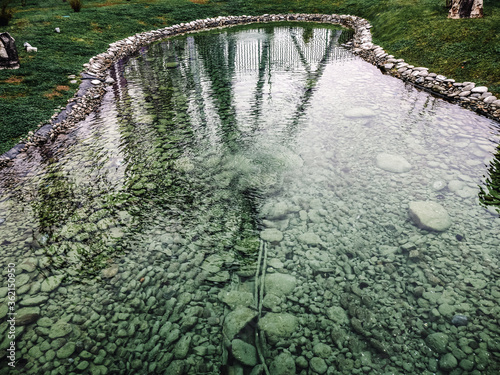 Transparent shallow pond with pebbles at the bottom with a rim of pebbles around the greenery and reflection of trees in the water