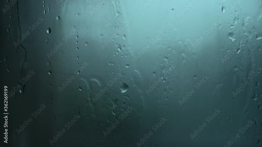 Close up view of rain drops trickling down on glass surface with blue background. Droplets of water running down during heavy rain. Close up view of rain drops trickling down on glass surface with blue background. Droplets of water running down during heavy rain.