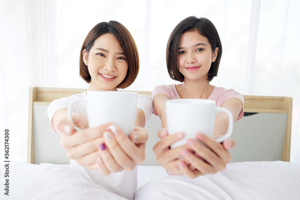 Asian young sisters lovely couple on white bed wake up and smiling with drinking cup of hot coffee together in bedroom. Homosexual women or Lesbian in love. Asian Teenage and Holiday concept.