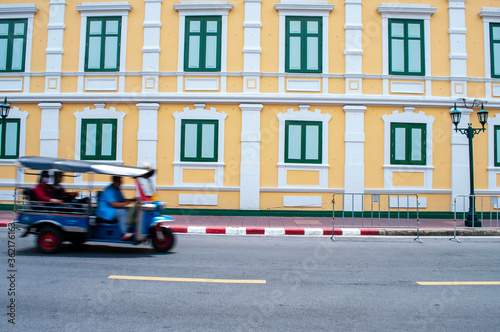tuk-tuk on the road in bangkok with yellow building background, speed motion.Moving Tuk tuk taxi with windows wall of The Ministry of Defence in Bangkok, Thailand © Albert