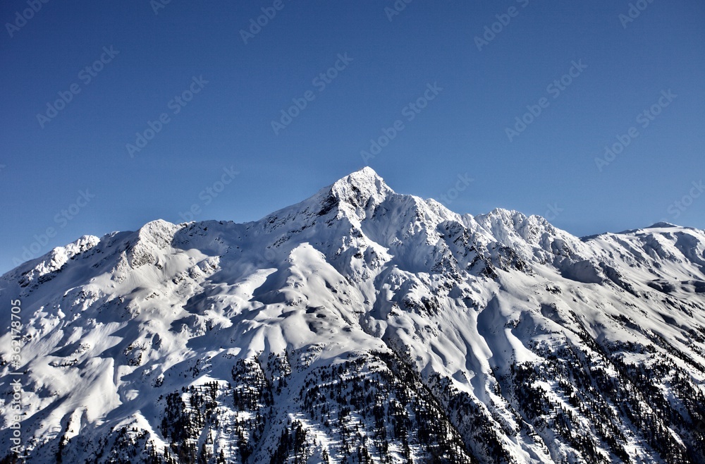White snowcapped isolated mountain peak on a sunny winter day. Snowy mountains winter landscape on bright clear blue sky background. Sunny snowy day in Alps mountains. Snow-covered mountain peak. 