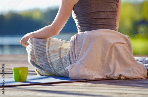 A woman is meditating on a wooden pier by the lake on a bright early sunny morning. Relaxing in lotus position after doing yoga outdoors. Relaxing tea ceremony and outdoor meditation. Happy life.