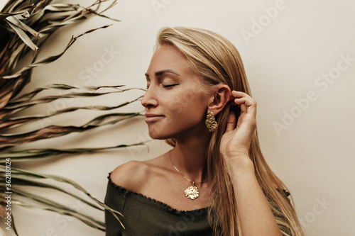 Tanned young blonde woman with gold earrings and medallion smiles with closed eyes. Pretty girl in khaki top poses near white wall with palm leaf. photo