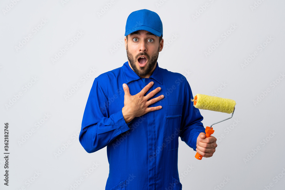 Painter man holding a paint roller isolated on white background surprised and shocked while looking right