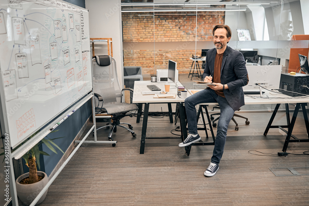 Smiling leader at company wearing jeans and jacket sitting on the table Smiling leader at company wearing jeans and jacket sitting on the table