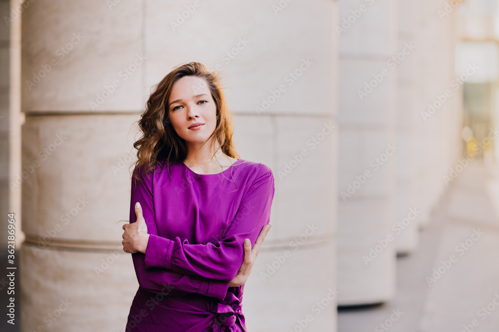 Attractive confident caucasian woman in violet dress hugging herself stands against blurry city background with free space. Mockup for ad. Business and financial people concept.