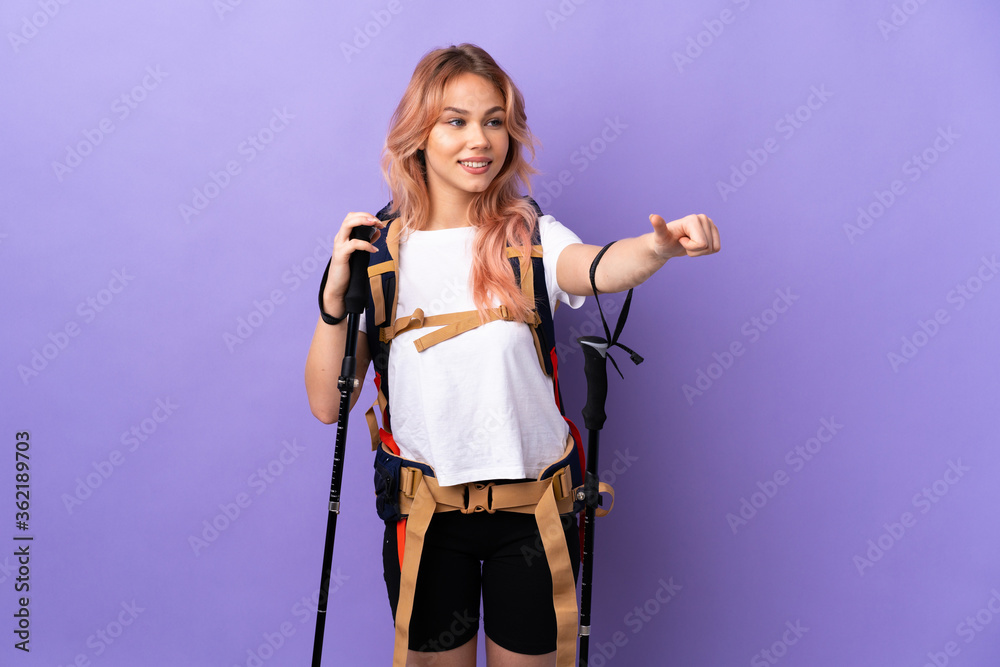 Teenager girl with backpack and trekking poles over isolated purple background giving a thumbs up gesture