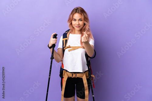 Teenager girl with backpack and trekking poles over isolated purple background making money gesture