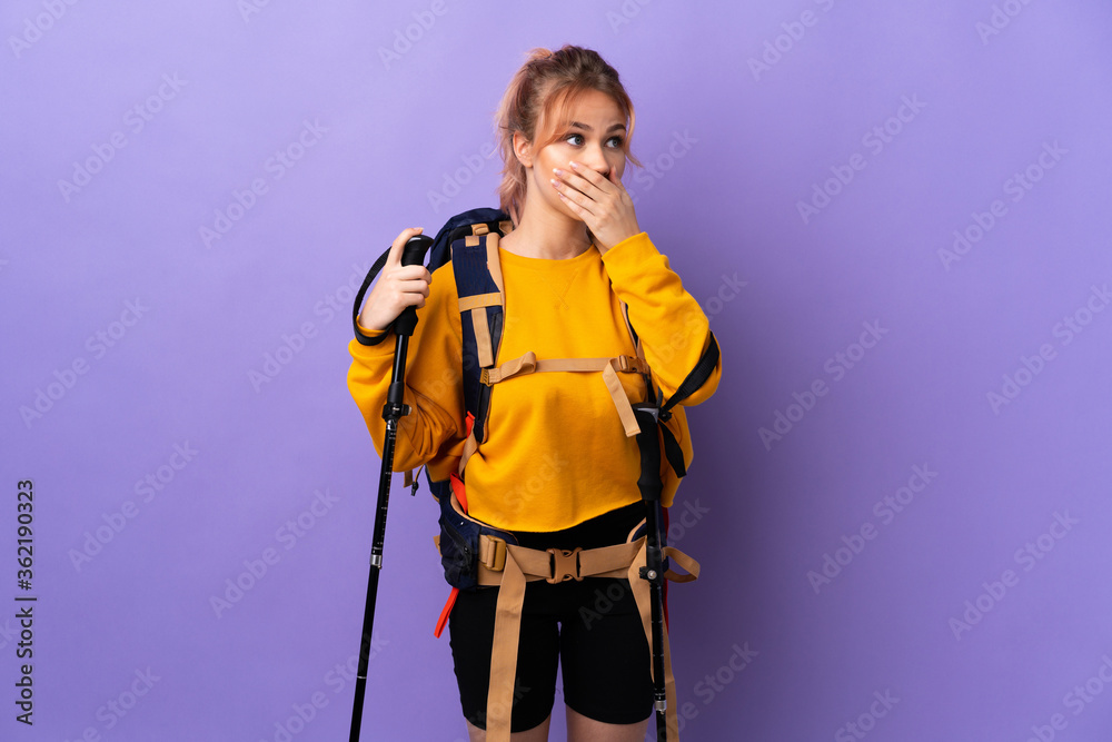 Teenager girl with backpack and trekking poles over isolated purple background covering mouth and looking to the side