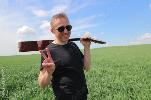 A man in black sunglasses stands in a field with a guitar on his shoulder. Happy guitar player in nature showing victory sign with fingers..
