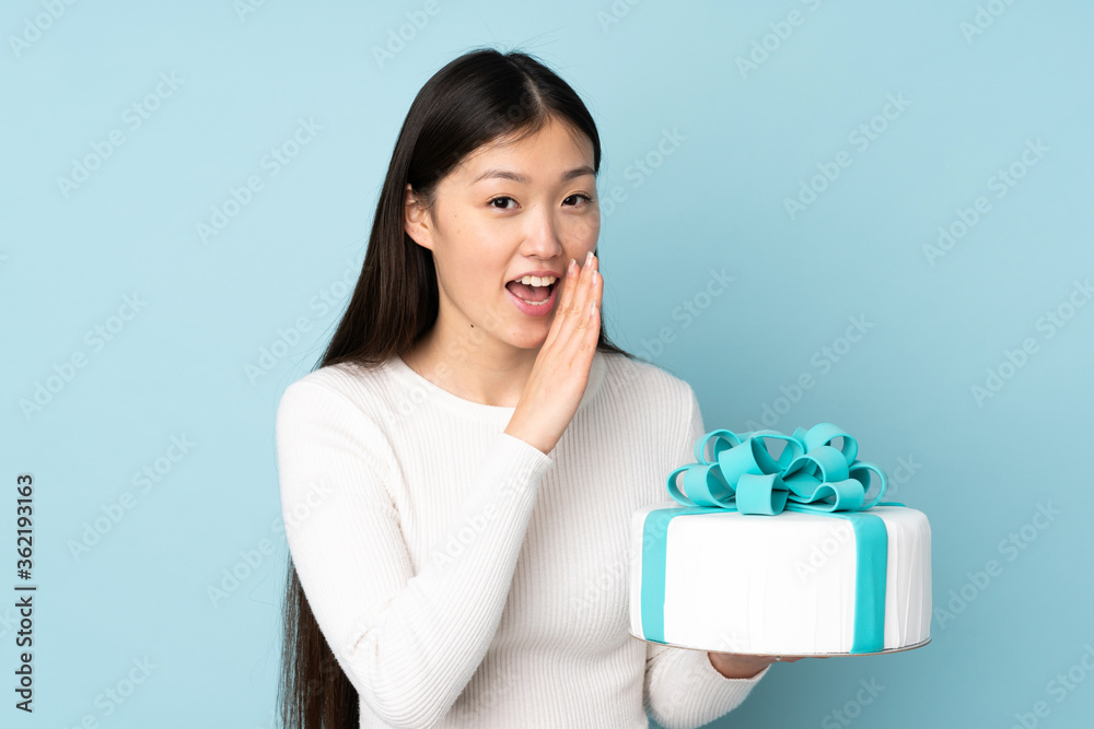 Pastry asian chef holding a big cake isolated on blue background whispering something Pastry asian chef holding a big cake isolated on blue background whispering something