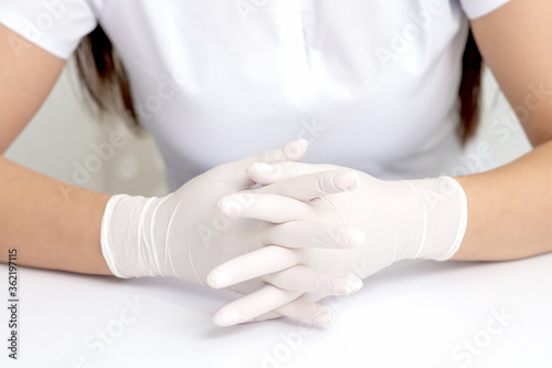 Hands in protective white medical gloves of the woman sitting at the white table. Crossed fingers in gloves