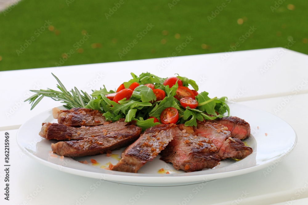 Juicy meat presented on a plate accompanied by a green salad and tomatoes on a beach table.
