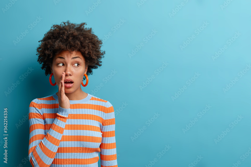 Mysterious woman with curly hair tells secret or confidential information, keeps hand near mouth and whispers gossips spreads rumors, looks aside, isolated on blue wall. Secrecy and hearsay concept - obrazy, fototapety, plakaty 