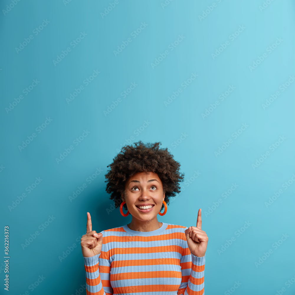 Cheerful African American woman looks up and points upwards with broad smile, recommends awesome product, suggests click banner, dressed casually, isolated on blue background shows advertisement promo - obrazy, fototapety, plakaty 