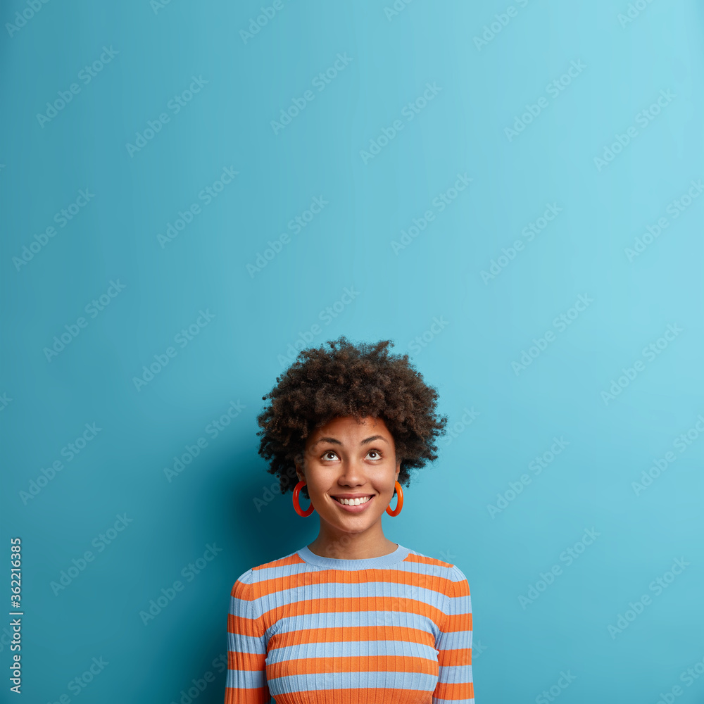 Vertical shot of good looking Afro American woman looks above with broad smile, notices nice offer or sale, feels happy dressed in casual striped jumper isolated on blue background. Copy space upwards - obrazy, fototapety, plakaty 