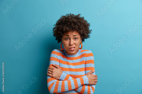 Photo of displeased curly woman hugs herself with crossed arms, needs to warm up in cold day, freezes on windy weather, frowns face and shakes from cold, wears only jumper without outerwear.