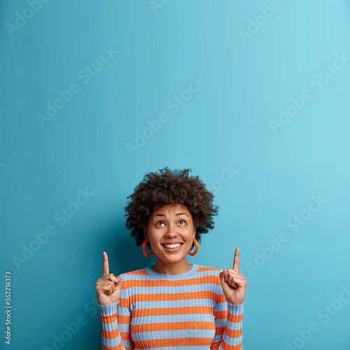 Cheerful African American woman looks up and points upwards with broad smile, recommends awesome product, suggests click banner, dressed casually, isolated on blue background shows advertisement promo