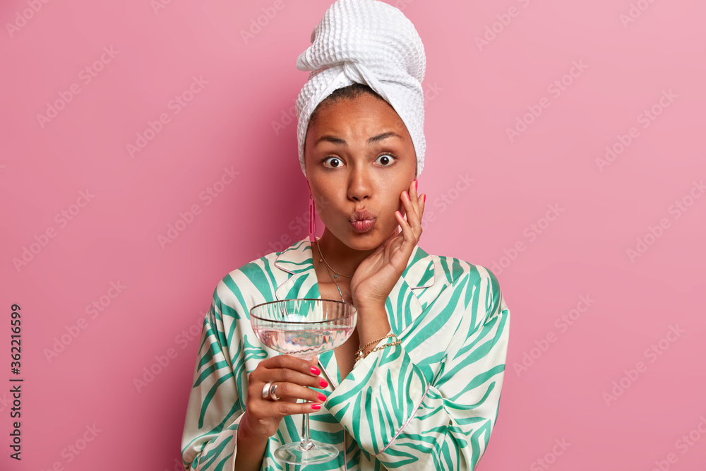 Well groomed young woman with healthy skin, keeps lips rounded, wears bath towel on head and dressing gown, drinks homemade cocktail, prepares for party at home, isolated on pink background. Well groomed young woman with healthy skin, keeps lips rounded, wears bath towel on head and dressing gown, drinks homemade cocktail, prepares for party at home, isolated on pink background.