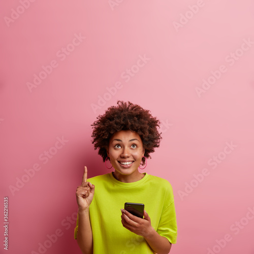 Amused cheerful African American woman holds modern mobile phone, connected to free internet, points upwards on blank space, demonstrates place for advertisement, smiles broadly, isolated on pink wall