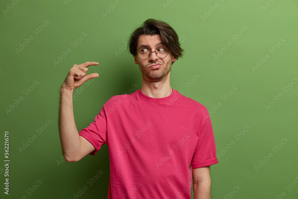 Displeased man asks some more time, shows small size or little bit gesture, needs minimum amount, measures not big scale, dressed casually, isolated on green background. Body language concept Displeased man asks some more time, shows small size or little bit gesture, needs minimum amount, measures not big scale, dressed casually, isolated on green background. Body language concept