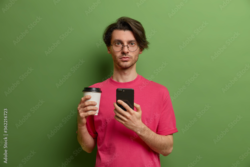 Serious man with trendy haircut exchanges messages via mobile phone, types and browses in network, drinks takeaway coffee, wears pink t shirt isolated on green background stands distracted from screen Serious man with trendy haircut exchanges messages via mobile phone, types and browses in network, drinks takeaway coffee, wears pink t shirt isolated on green background stands distracted from screen