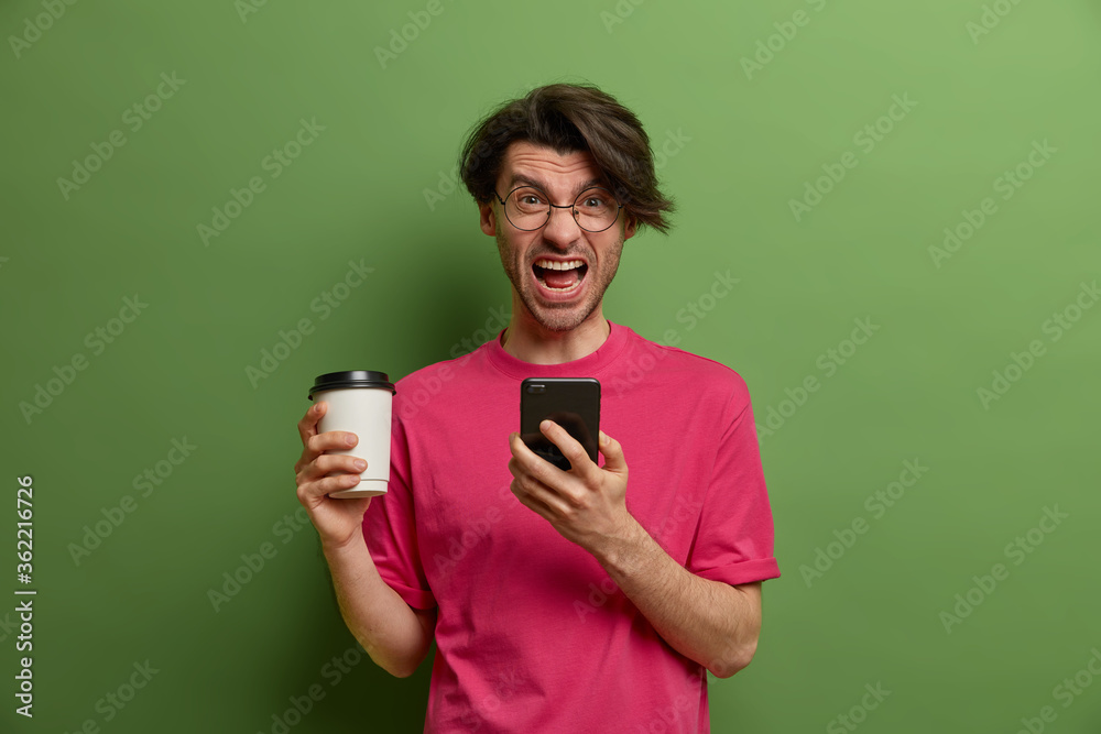 Crazy man shouts loudly, holds coffee in paper cup, uses modern smartphone, surfs web, annoyed to get bad message, dressed in pink t shirt, dissatisfied after talking with boring interloctor Crazy man shouts loudly, holds coffee in paper cup, uses modern smartphone, surfs web, annoyed to get bad message, dressed in pink t shirt, dissatisfied after talking with boring interloctor