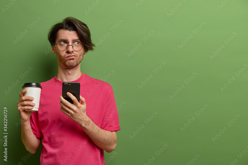 Modern lifestyle and technology concept. Pensive serious man with dark hair uses app or plays game, thinks about something while drinks coffee from paper cup, stands against green wall, blank space Modern lifestyle and technology concept. Pensive serious man with dark hair uses app or plays game, thinks about something while drinks coffee from paper cup, stands against green wall, blank space