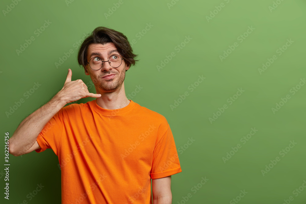 Call me! Dreamy young Caucasian man makes telephone hand gesture, offers to contact by phone, wears round transparent glasses and orange t shirt, isolated on green background, blank copy space Call me! Dreamy young Caucasian man makes telephone hand gesture, offers to contact by phone, wears round transparent glasses and orange t shirt, isolated on green background, blank copy space