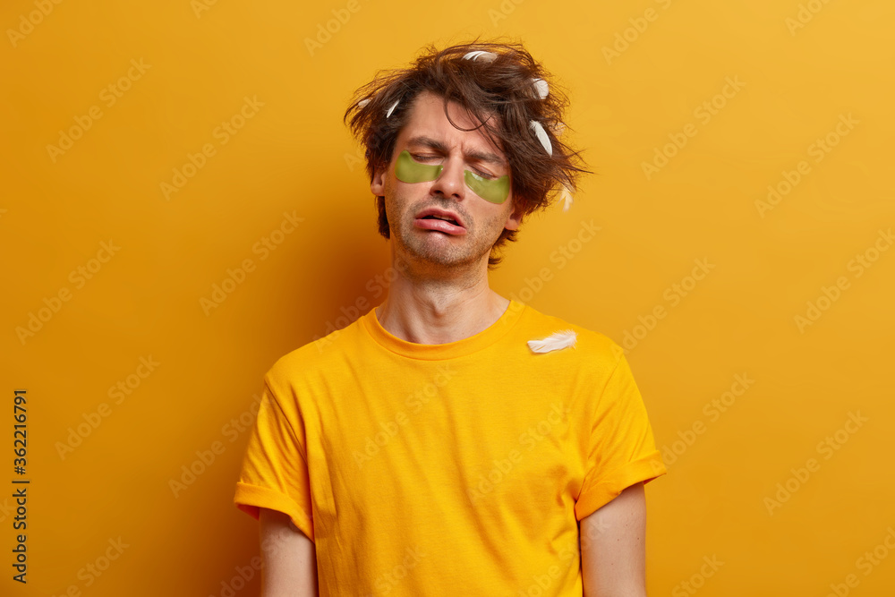 Exhausted tired sleepy young man cannot awake very early, prepares for important day, wears collagen patches for reducing puffiness under eyes, dressed in yellow casual clothes. People and tiredness Exhausted tired sleepy young man cannot awake very early, prepares for important day, wears collagen patches for reducing puffiness under eyes, dressed in yellow casual clothes. People and tiredness