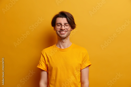 Joyful positive man closes eyes and smiles pleasantly, poses with anticipation of something good happen, dressed in bright yellow t shirt, stands indoor, expresses optimism. Monochrome shot.