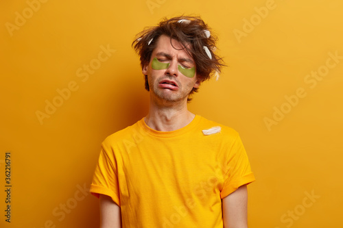 Exhausted tired sleepy young man cannot awake very early, prepares for important day, wears collagen patches for reducing puffiness under eyes, dressed in yellow casual clothes. People and tiredness © Wayhome Studio