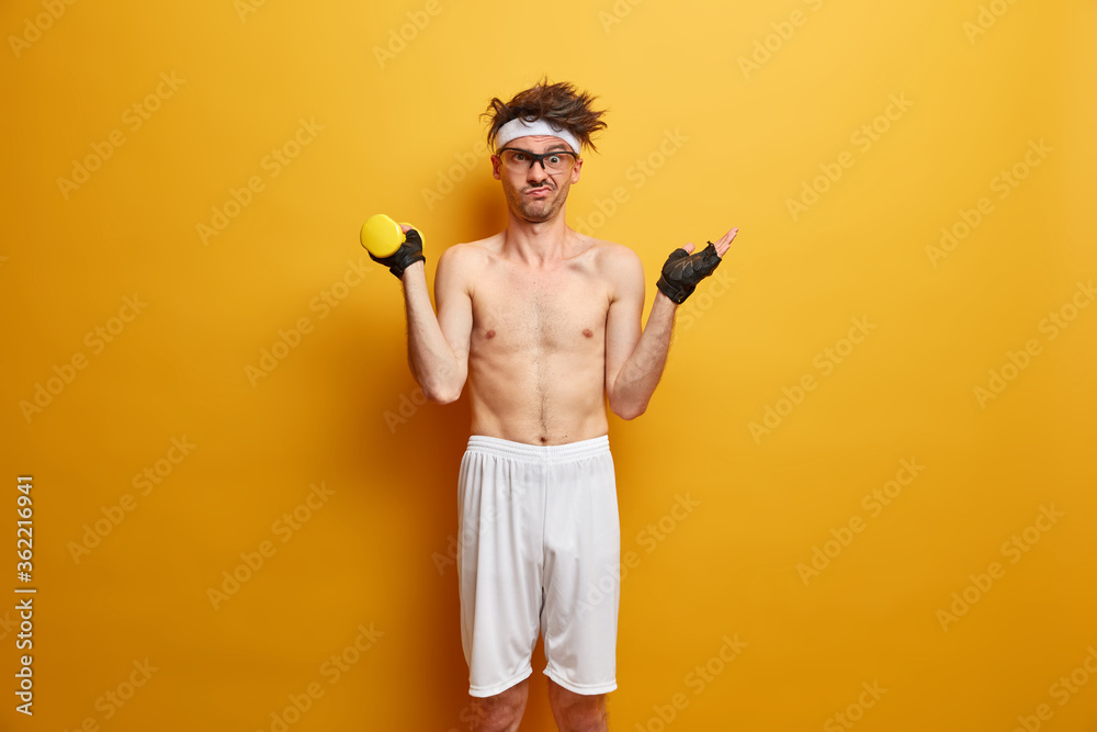 Healthy lifestyle, fitness and bodybuilding concept. Puzzled man raises hand in bewilderment, does workout at gym, lifts dumbbell, poses shirtless against yellow background, doesnt like sport Healthy lifestyle, fitness and bodybuilding concept. Puzzled man raises hand in bewilderment, does workout at gym, lifts dumbbell, poses shirtless against yellow background, doesnt like sport