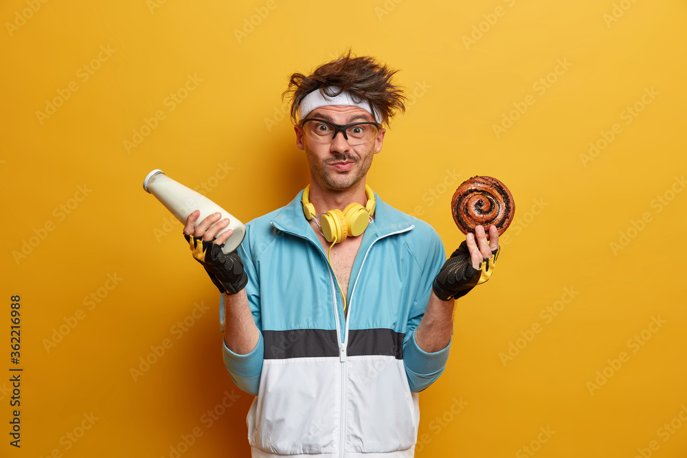 Sportsman poses with bottle of milk and sweet bun, asks not to eat junk food for keeping healthy lifestyle, wears sport clothes, transparent glasses, isolated on yellow background. Sport and nutrition