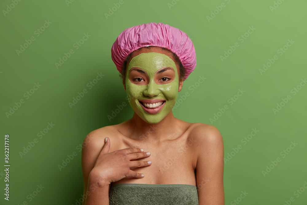 Happy ethnic woman spends free time in spa salon, applies cosmetic mask on face, wears bath hat on head, stands with bare shoulders against green background. Beauty treatment and skin care concept Happy ethnic woman spends free time in spa salon, applies cosmetic mask on face, wears bath hat on head, stands with bare shoulders against green background. Beauty treatment and skin care concept