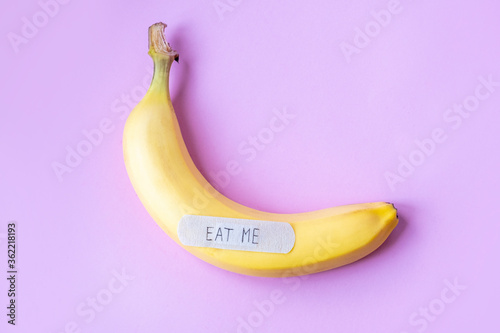 Yellow banana with plaster note EAT ME on pastel pink background. Minimal food concept. Flat lay, top view.