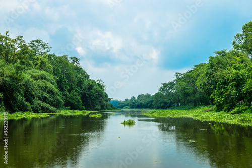 A landscape of a river with transparent water flowing through a village with surrounding greenery. Blue sky above with its reflection on the water