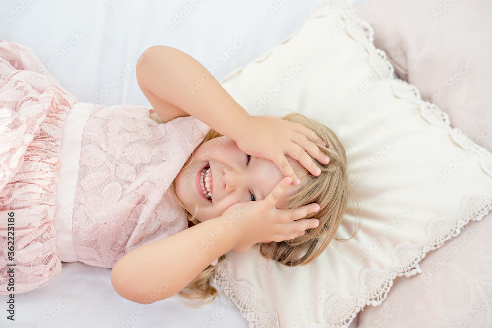 Beautiful little blonde girl with blue eyes. A close up portrait, the baby lies on white sheets with pillows of a bed with chiffon canopies against the blue sea. Happy childhood and summer vacation Beautiful little blonde girl with blue eyes. A close up portrait, the baby lies on white sheets with pillows of a bed with chiffon canopies against the blue sea. Happy childhood and summer vacation