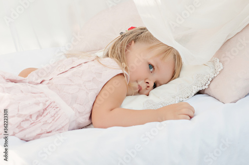 Beautiful little blonde girl with blue eyes. A close up portrait, the baby lies on white sheets with pillows of a bed with chiffon canopies against the blue sea. Happy childhood and summer vacation