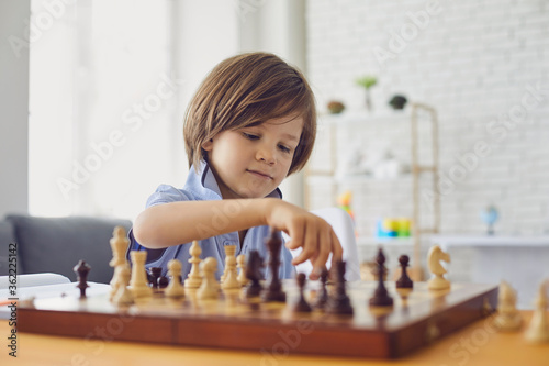 Smart boy plays chess at the table in the room. The child plays board games.