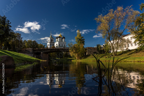 View of the Pskov Kremlin over the bridge at the city river on a summer clear day. The blue bright sky with clouds is reflected in a smooth surface of water.
