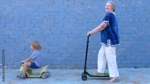 Funny blond boy in plaid shirt riding bicycle with his beloved grandmother. child teaches an elderly granny to ride. pensioner is smartly dressed and rolls on a large stretched scooter
