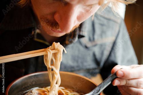 white blonde hipster man looking down to eat and hot slurp japanese buckwheat noodle soba with chopsticks