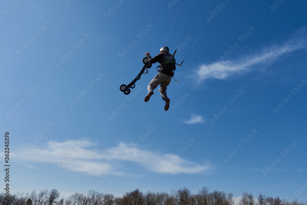 Man jumps through the air, holding in his hand an all terain board, tree line and blue sky with white clouds. Germany.