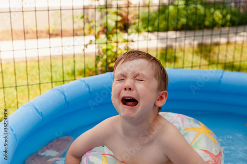 Little unhappy crying boy sitting in the blue inflatable pool on the backyard.