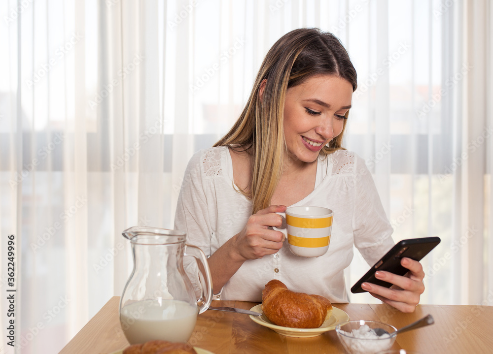 Pretty young blonde woman using phone while eating breakfast at home
