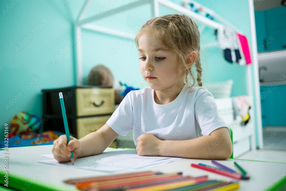 Little girl draws while sitting at a table in a room against the background of the wall