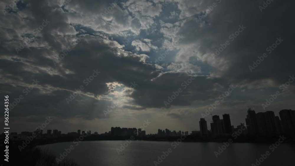 Time-lapse, dark clouds floating over city and lake, sunlight through thick clouds, light beam