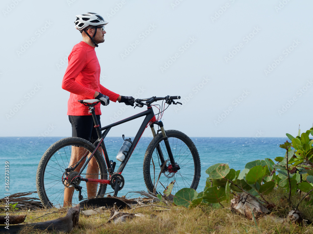 Obraz premium Fitness man with a bicycle stands on the ocean shore and looks into the distance on a sunny day. Athletic healthy people active lifestyle during pandemic coronavirus..